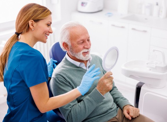 Man smiling while looking at reflection in handheld mirror with dentist
